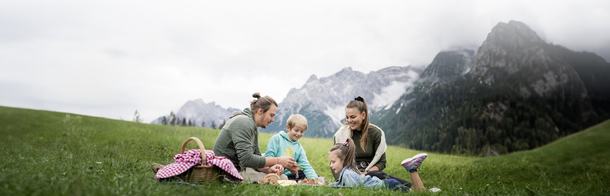 Alpine Stays for all Generations by Family Rainer Familie macht Picknick auf einer grünen Wiese mit Bergen im Hintergrund