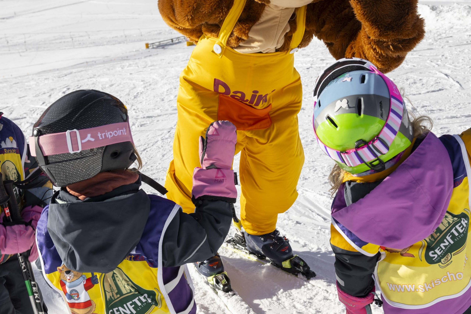 Experience exciting adventures with Raini! Children on skis high-five a mascot in yellow pants on the snow