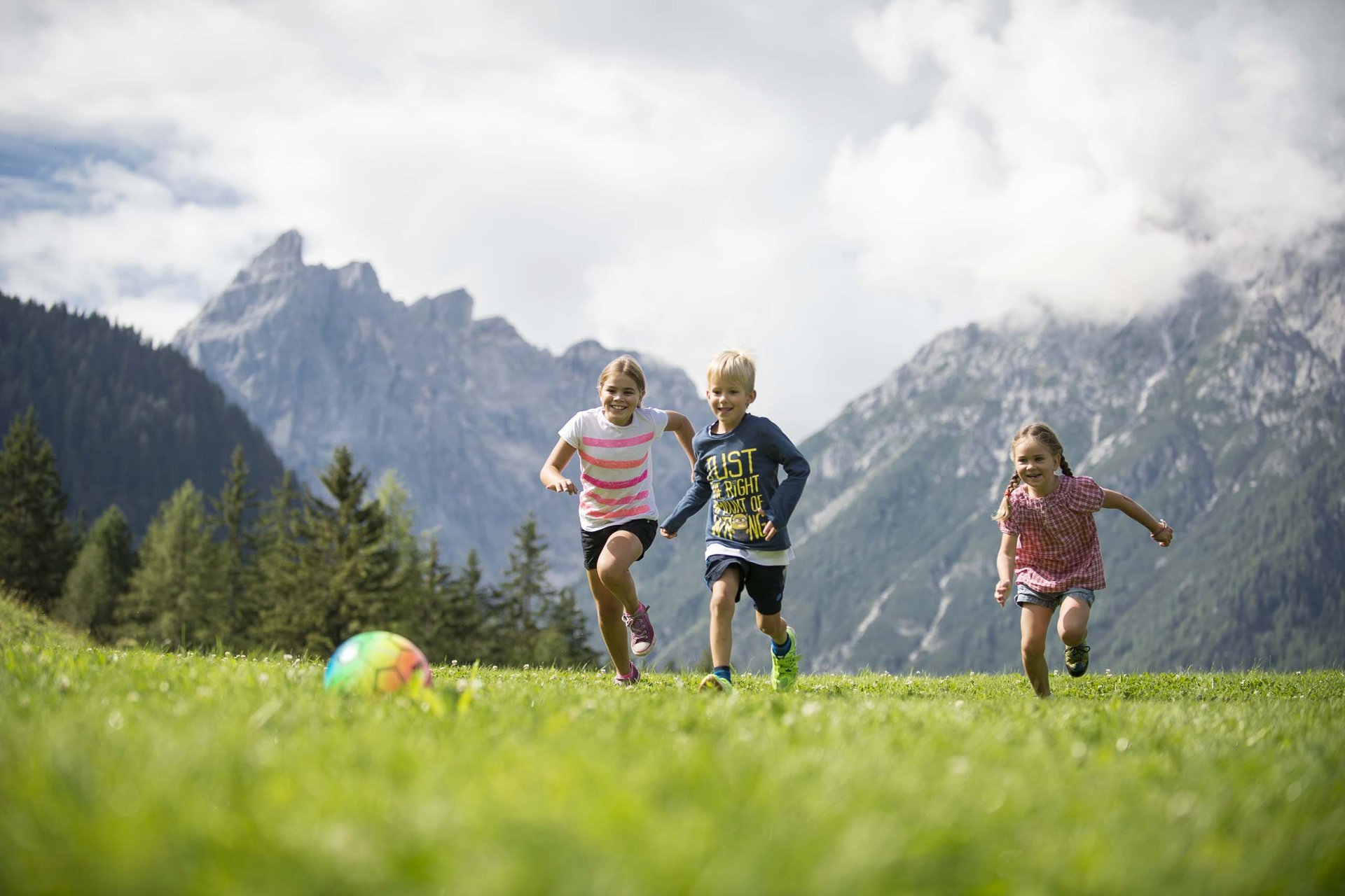 Experience exciting adventures with Raini! Three children playing with colorful ball on grass in front of mountains