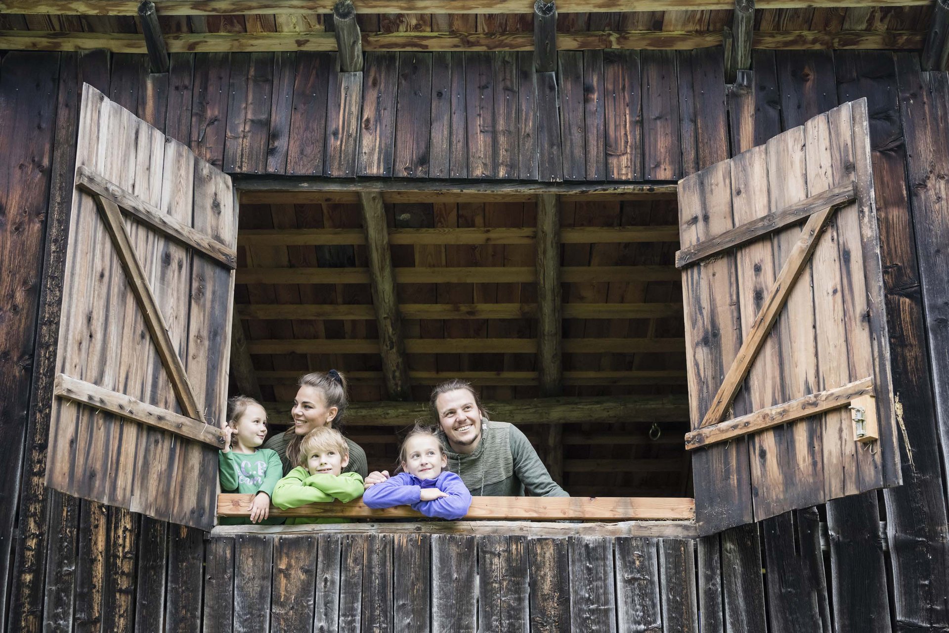 Your family resort in the Dolomites Family looking out of open wooden window in rustic barn