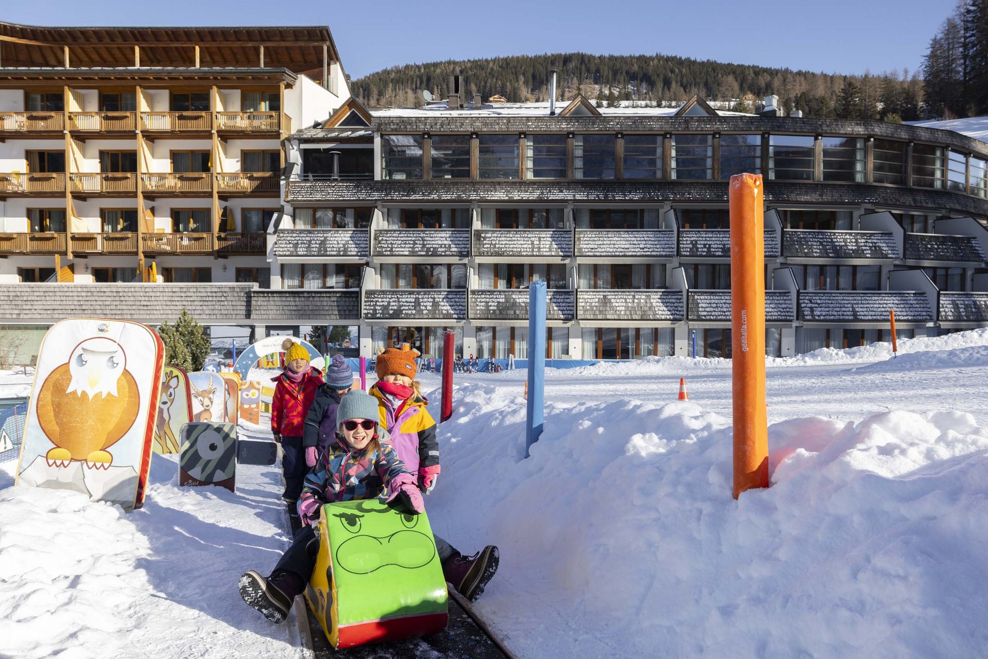 Experience exciting adventures with Raini! Children sledding on a snowy path in front of a hotel in winter
