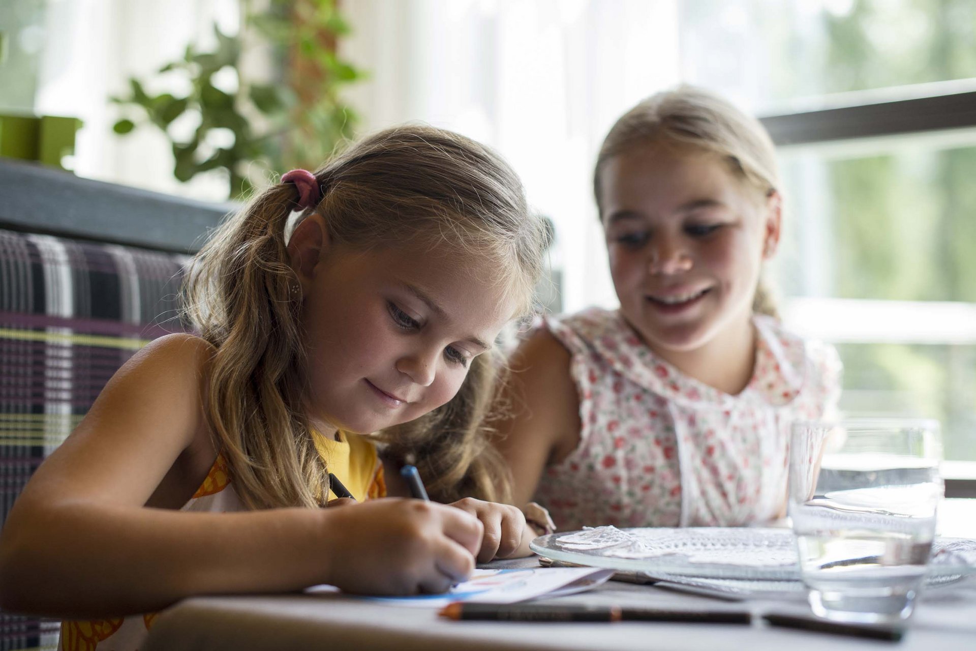 Experience exciting adventures with Raini! Two girls sitting at a table, one drawing focused