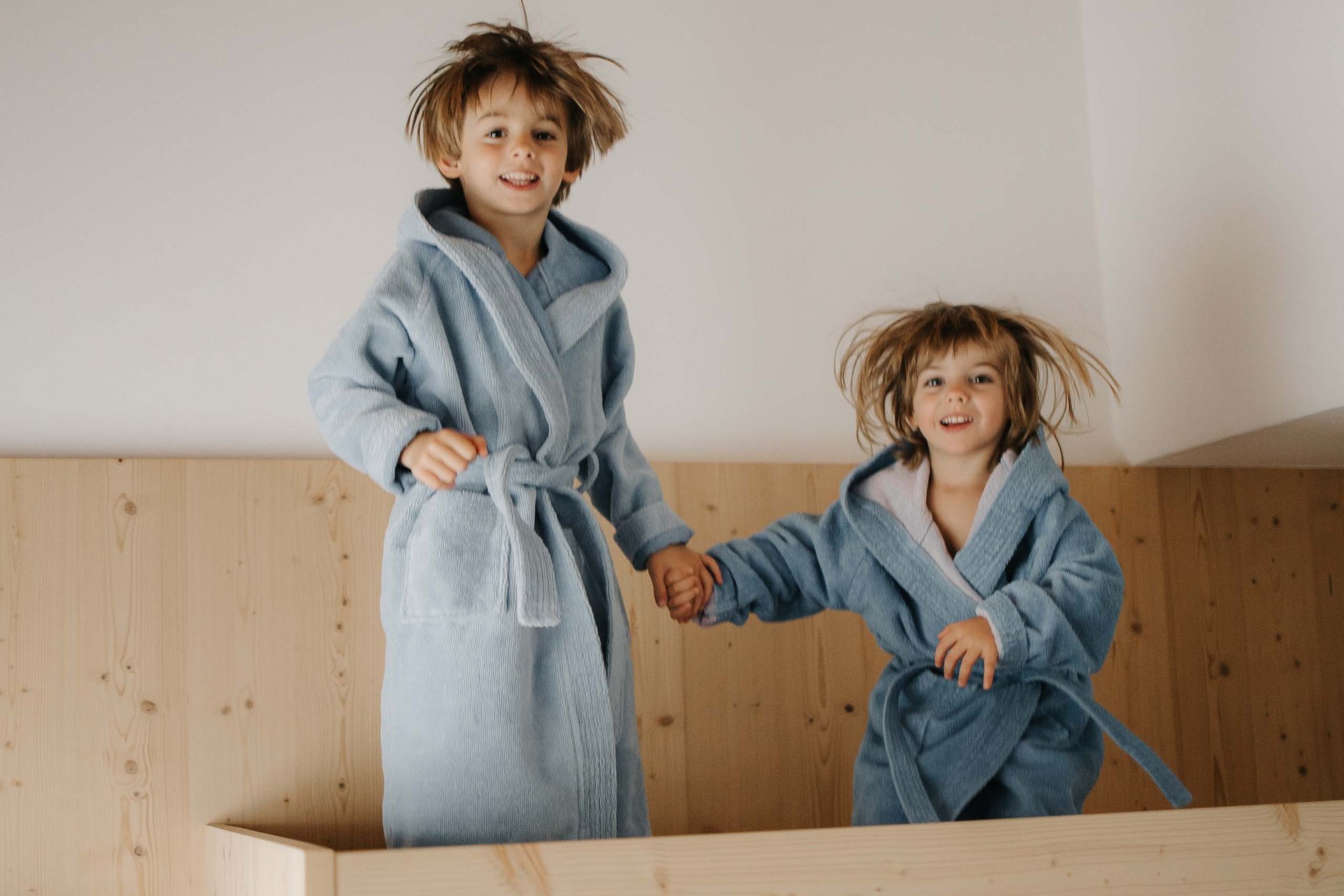 Your family resort in the Dolomites Two children in blue bathrobes happily jumping on a wooden bed