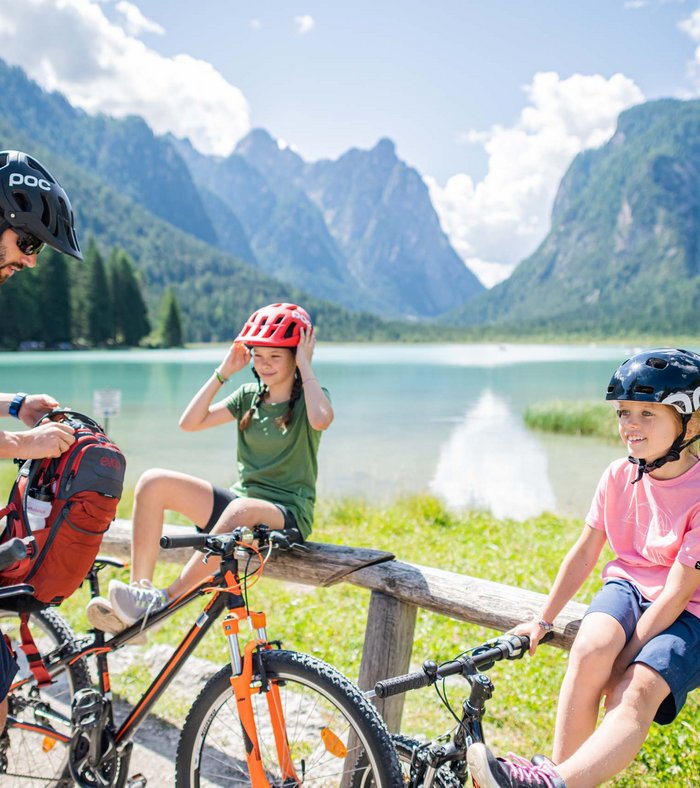 True adventure at our family hotel in the Dolomites! Family with bikes taking a break at alpine mountain lake on a sunny day
