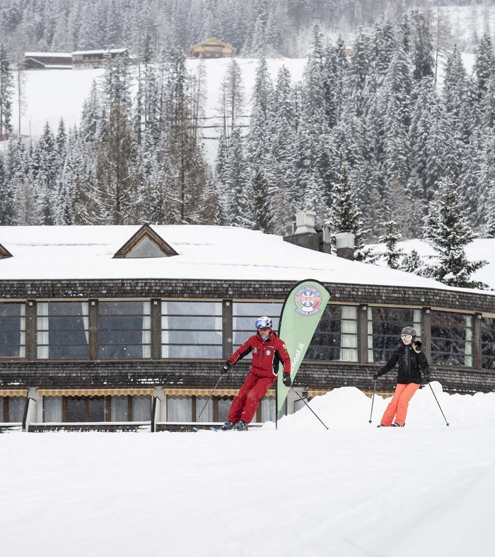 1 Familienhotel in Südtirol und 3 Residences: Family Resort Rainer Skilehrer gibt Skistunde vor verschneitem Gebäude und Wald