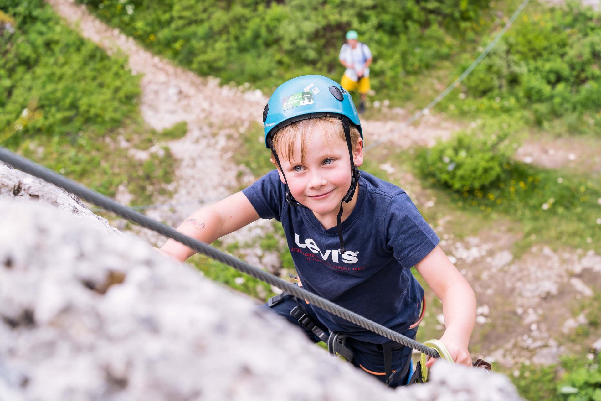 Your family resort in the Dolomites Boy wearing helmet climbs rock face, person belays below on green terrain