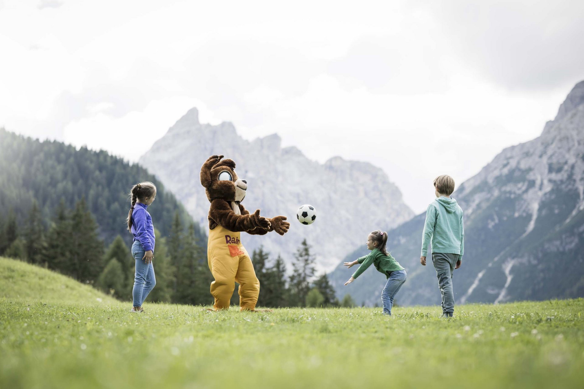 Experience exciting adventures with Raini! Children play with a person in a bear costume on a grassy field with mountains