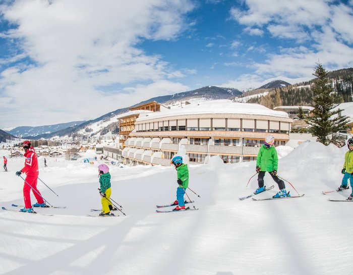 Alpine Stays for all Generations by Family Rainer Skikurs mit Kindern und Lehrer vor einem Hotel in verschneiter Berglandschaft