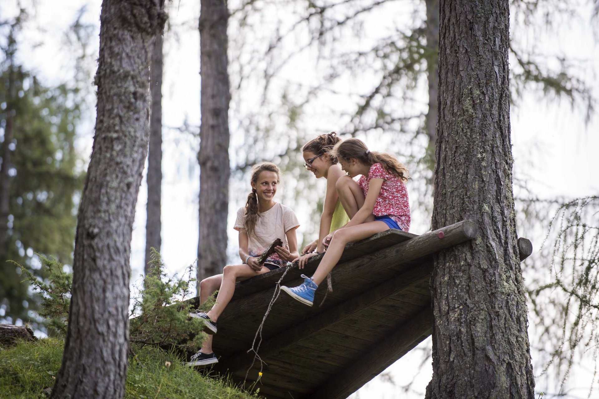 Experience exciting adventures with Raini! Three girls sitting and laughing on a wooden structure in the forest