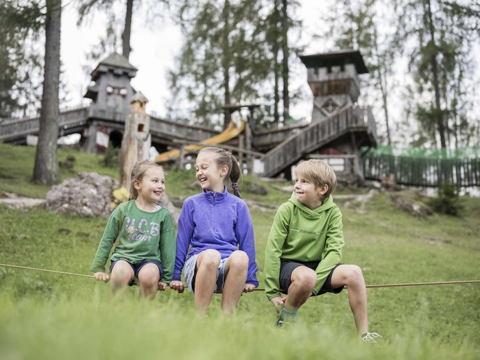 Experience exciting adventures with Raini! Three smiling children sitting on a rope in front of a wooden playground outdoors