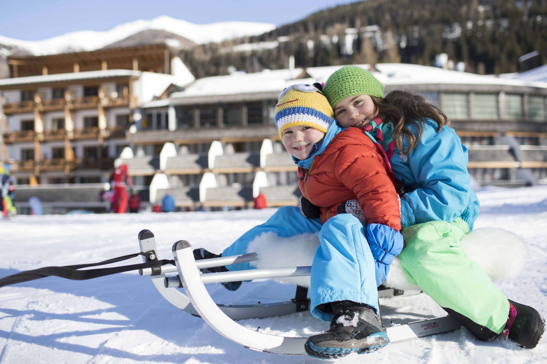 Experience exciting adventures with Raini! Two children sledding on snow in front of a hotel