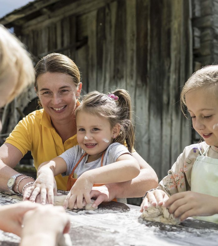 Alpine Stays for all Generations by Family Rainer Frau hilft Kindern beim Kneten von Teig draußen am Holztisch