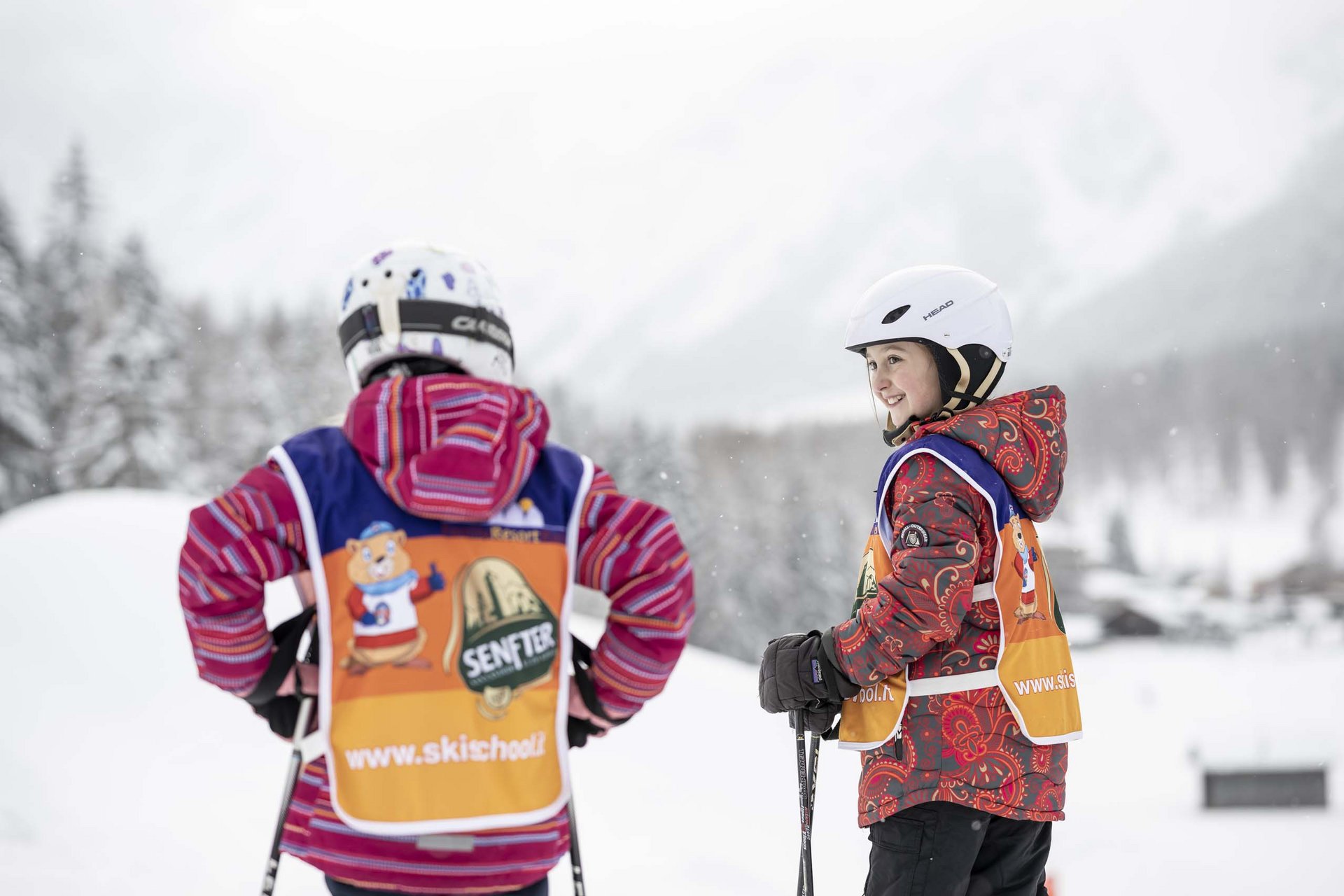 Experience exciting adventures with Raini! Two children in ski class wearing helmets and holding ski poles in the snow